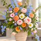 A vibrant floral arrangement in a beige ribbed ceramic pot, featuring peach roses, pink carnations, blue delphiniums, and green foliage, set on a light stone surface with a softly blurred indoor background.