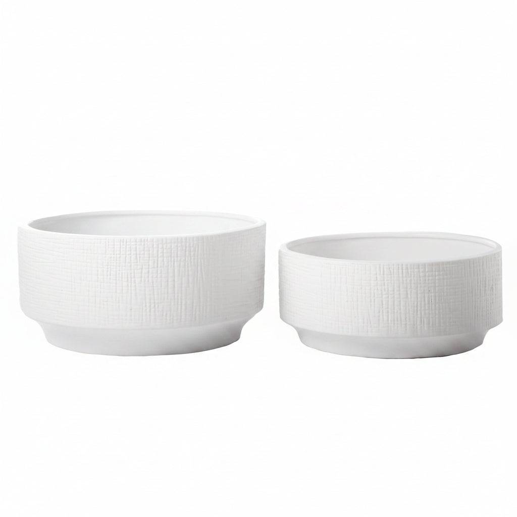Two white ceramic round planters of different sizes placed on a surface with a blurred background featuring a plant.