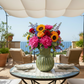 Colorful flower arrangement in a green vase on a table with chairs and an umbrella in the background.