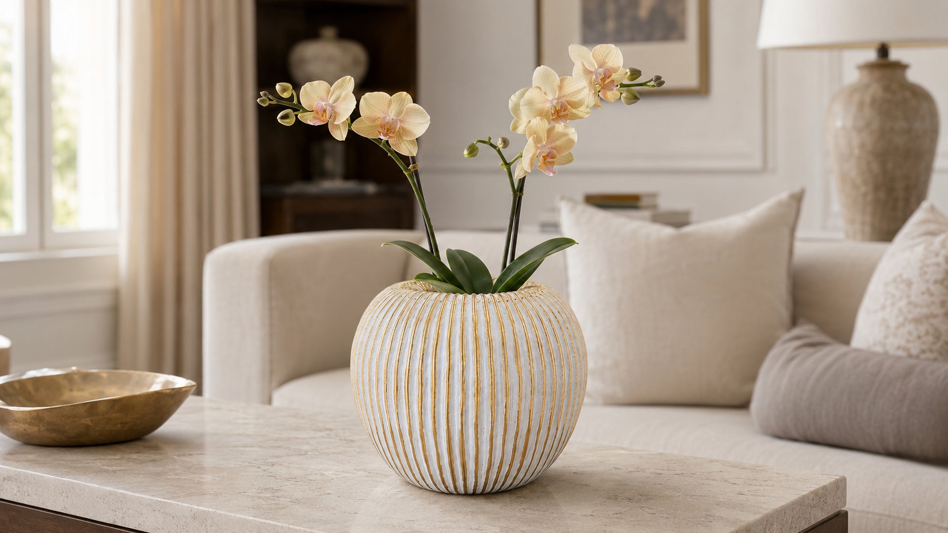 A ribbed ceramic vase with a warm cream and gold finish, filled with pale yellow orchids, set on a marble coffee table in a beautifully styled living room.