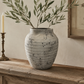 A textured, speckled gray ceramic vase filled with olive branches, displayed on a rustic wooden console table in a warm, neutral-toned room next to a candlestick and framed botanical art.