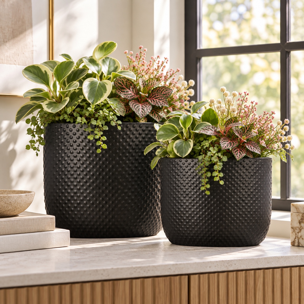 Two black textured planter pots filled with green and variegated houseplants, displayed on a light stone countertop near a window.