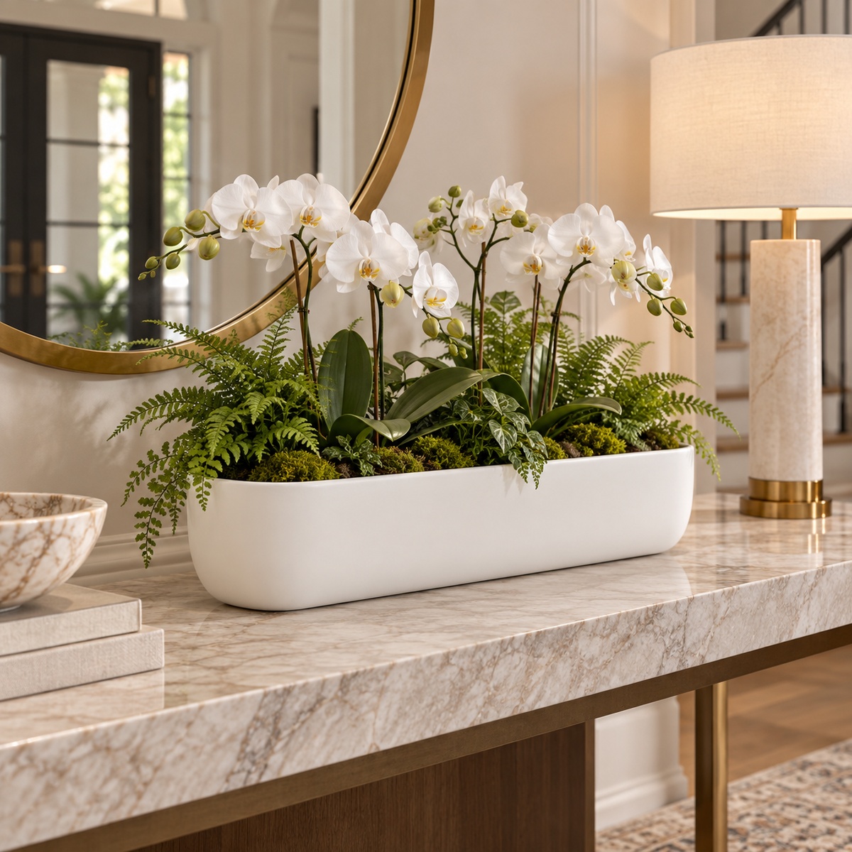 A long white rectangular planter filled with white orchids and green ferns, displayed on a marble console table in a bright, elegant interior setting.