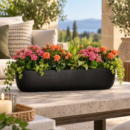 A long rectangular black planter filled with pink flowers, orange roses, and green foliage, placed on a stone patio table with an outdoor garden background.
