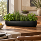 A long black rectangular planter filled with fresh green herbs, including basil, rosemary, and mint, displayed on a wooden dining table in a warm, modern kitchen setting.