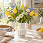 A white ribbed ceramic vase filled with a spring flower bouquet of yellow tulips, white daisies, blue delphiniums, and white lisianthus, displayed on a marble dining table in a bright, sunlit kitchen.