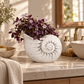 A white textured nautilus shell planter filled with dark purple and light purple flowers, styled on a beige marble bathroom counter.