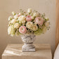 A floral arrangement of pale pink and cream flowers in a distressed white ceramic urn, displayed on a light wood bedside table next to a bed.