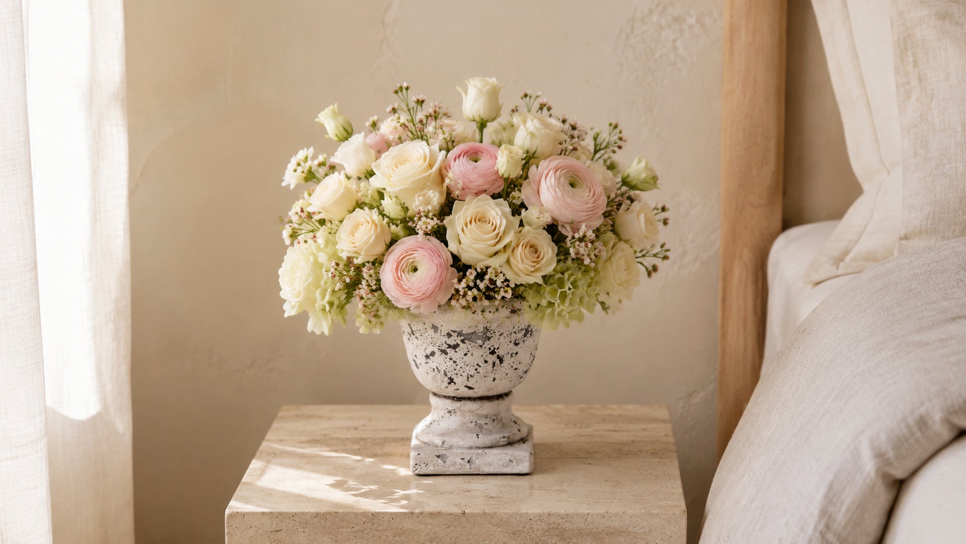 A floral arrangement of pale pink and cream flowers in a distressed white ceramic urn, displayed on a light wood bedside table next to a bed.