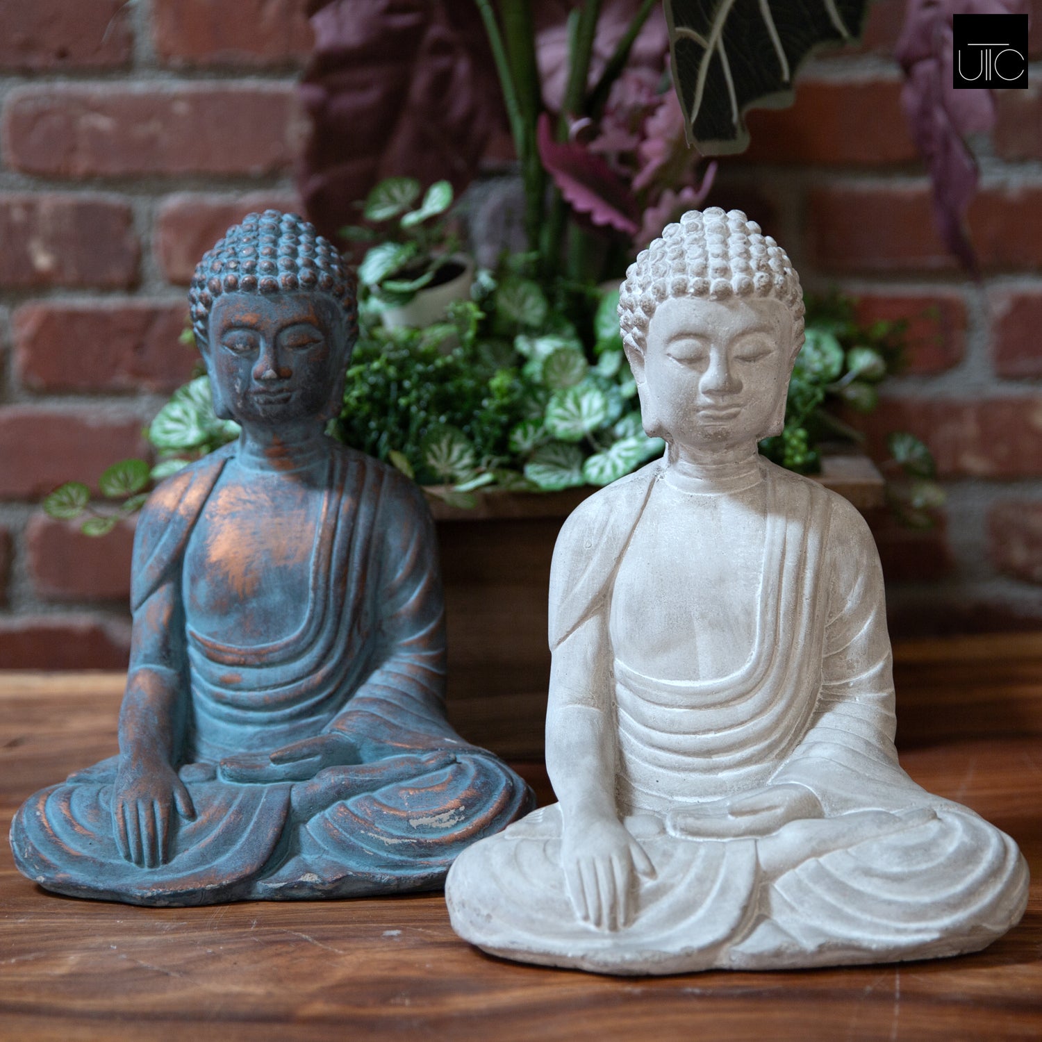 Two cement meditating Buddha figurines with a washed concrete finish, one in blue and one in gray, displayed against a brick backdrop with greenery.