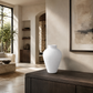 A textured white ceramic vase with a wide bulbous body and narrow neck, displayed on a dark wood console table in a modern, sunlit minimalist living room.