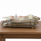 Three stacked rectangular gold-toned decorative trays with ornate openwork edges and small feet, styled on a wooden table with a glass bottle, pearl strand, pink feather, and candle.