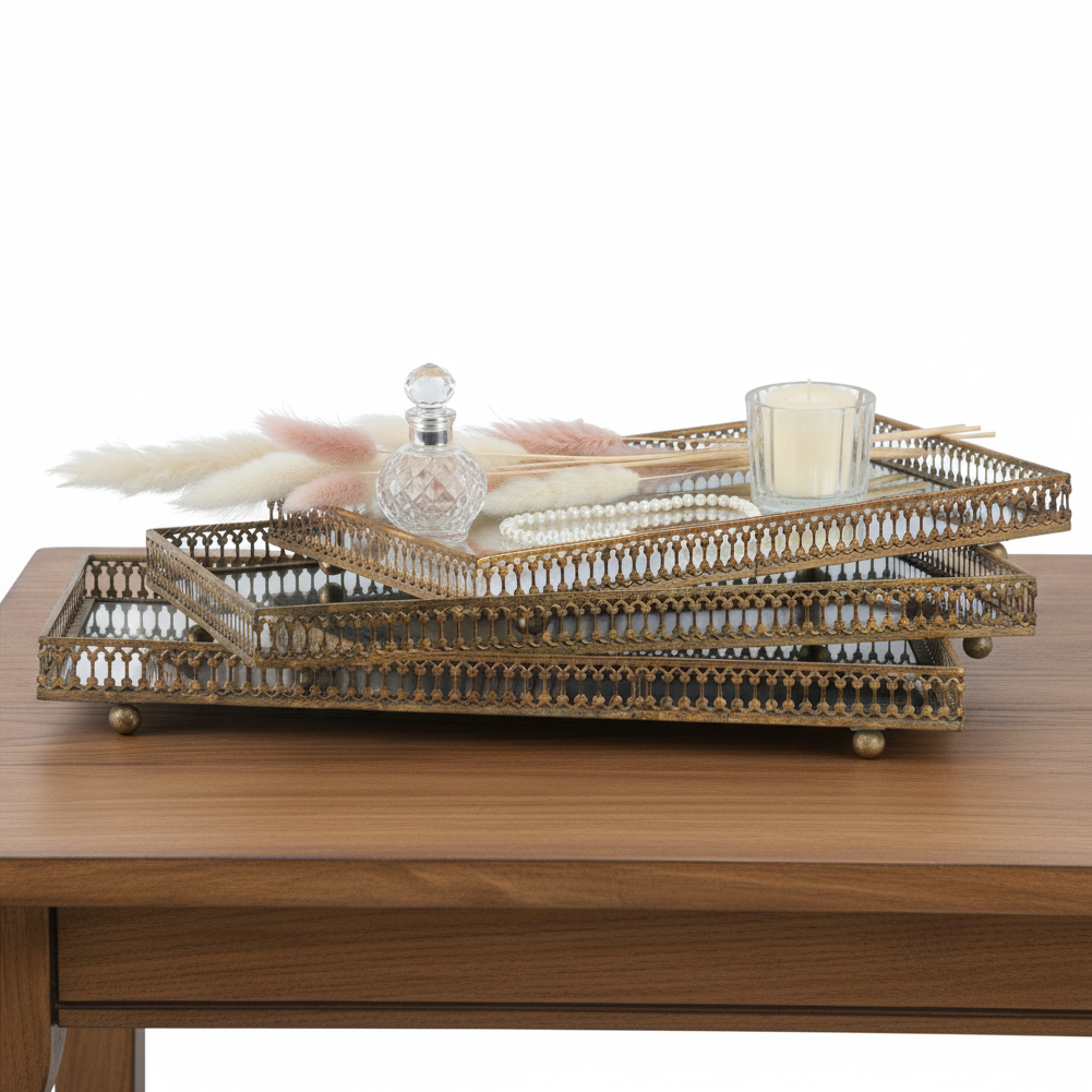 Three stacked rectangular gold-toned decorative trays with ornate openwork edges and small feet, styled on a wooden table with a glass bottle, pearl strand, pink feather, and candle.