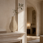 A minimalist beige ceramic vase with a single leafy sprig, displayed on a stone fireplace mantel in a warm, elegant room with an arched doorway and a sofa in the background.