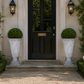 A black front door set in a beige stucco entryway, flanked by two large white stone planters with rounded green shrubs, with ivy climbing the walls and white flowers at the base.