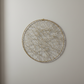 A circular wall hanging with a thin gold-toned wire frame and dense intersecting string webbing, mounted on a plain light gray wall.