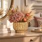 A decorative dried floral arrangement in a textured gold ceramic vase, styled on a wooden dresser in a warm, softly lit bedroom or living space with pillows, a mirror, and books in the background.