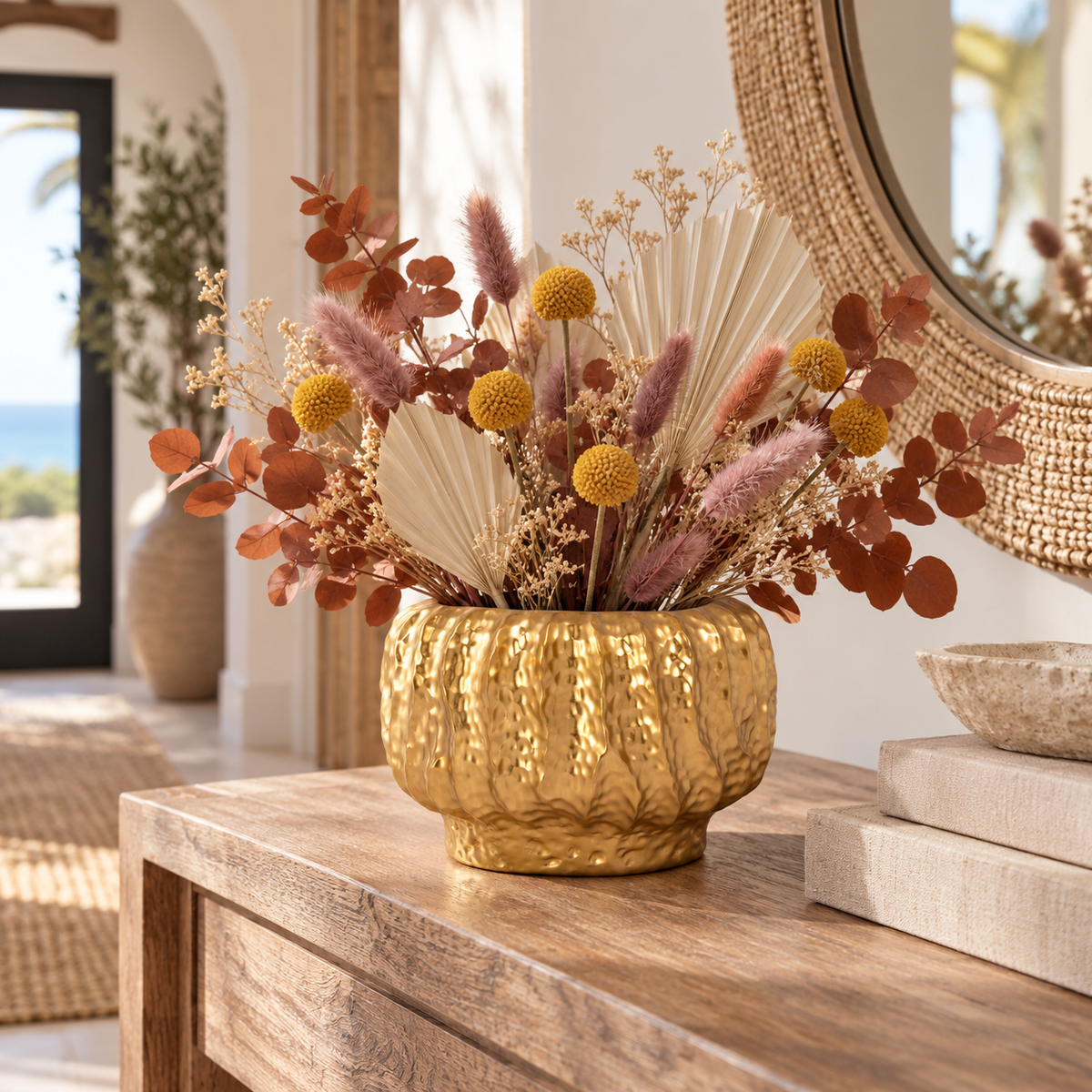 A textured gold ceramic vase filled with a dried floral arrangement featuring beige palm fronds, dusty pink bunny tails, and orange-brown eucalyptus, displayed on a wooden console table in a bright, modern interior.