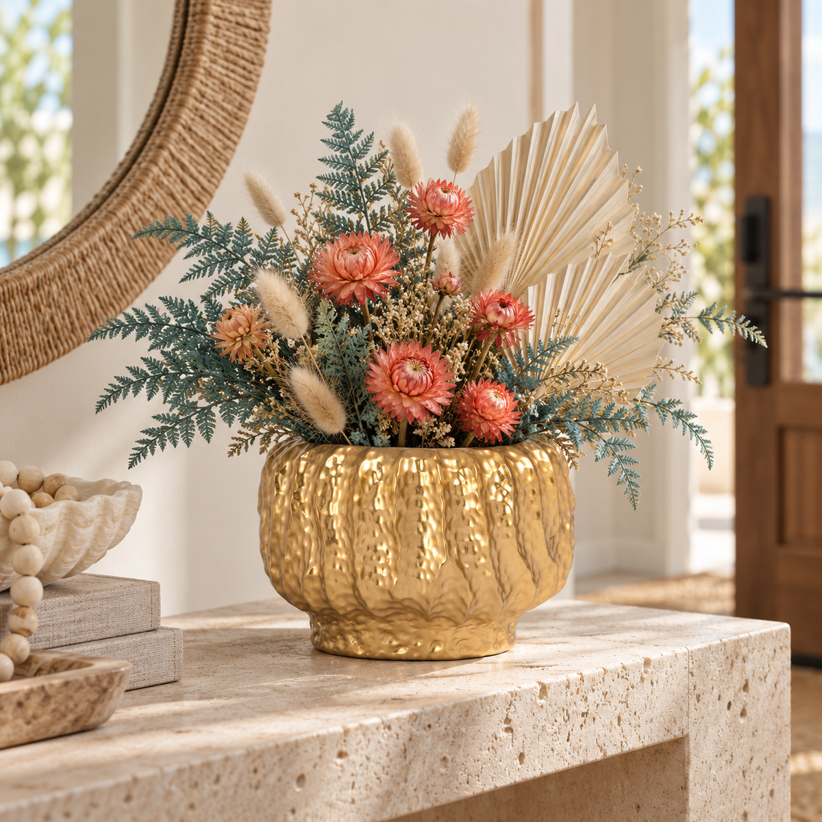 A decorative dried floral arrangement in a textured metallic gold pot, styled on a stone console table in a bright, modern interior setting.