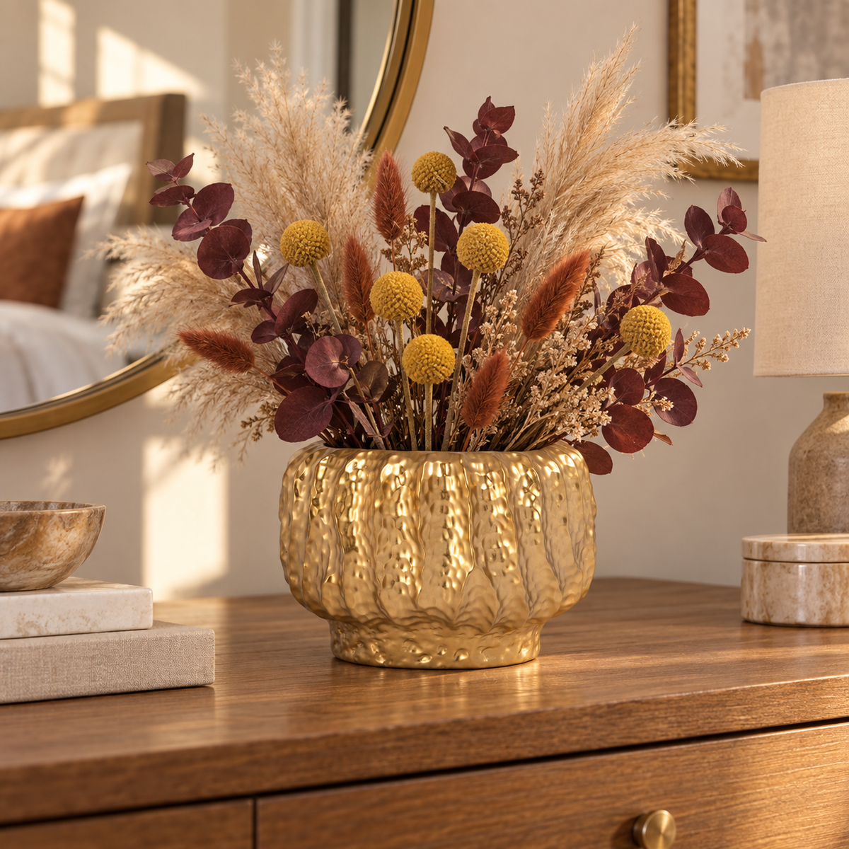 A decorative dried floral arrangement in a textured metallic gold vase, displayed on a wooden dresser in a warm, sunlit bedroom setting.