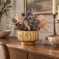 A decorative dried floral arrangement in a textured gold vase, displayed on a wooden console table in a styled interior setting with candles and bowls.