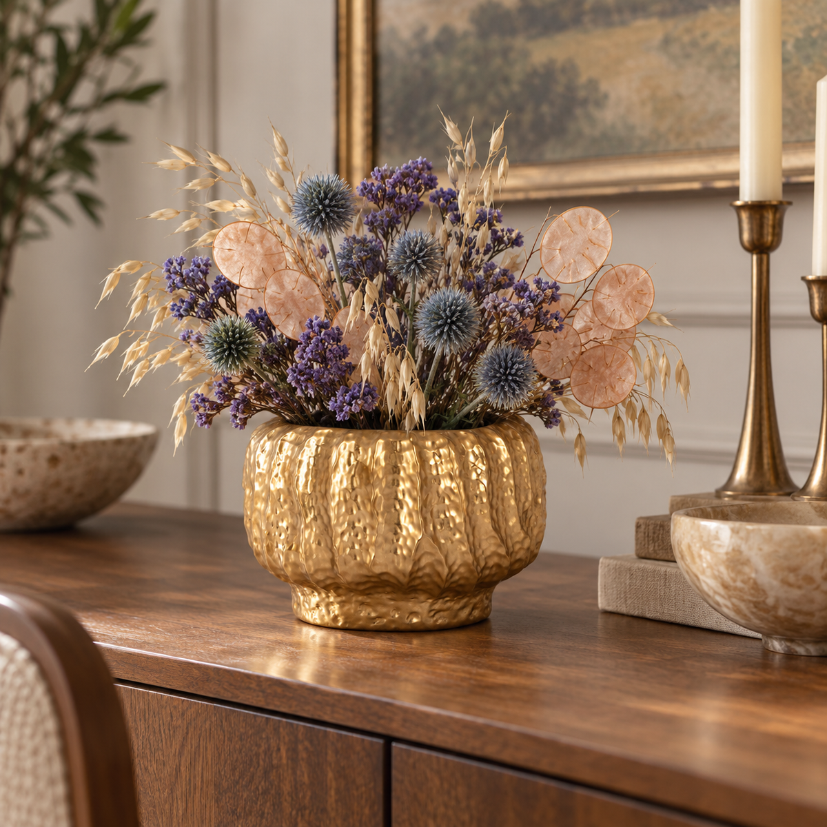 A decorative dried floral arrangement in a textured gold vase, displayed on a wooden console table in a styled interior setting with candles and bowls.