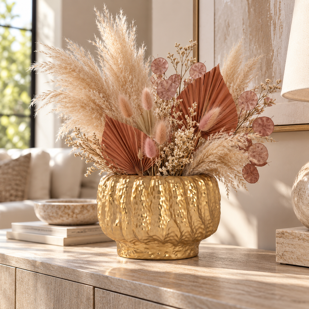 A decorative dried floral arrangement in a textured gold ceramic vase, styled on a light wood console table in a bright, modern living room.