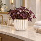 A white ceramic planter with vertical gold stripes sits on a marble bathroom counter, filled with deep burgundy foliage and small light pink flowers. A gold faucet, soap dispenser, mirror, and folded white towels are visible in the softly blurred background.