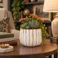 A white ceramic planter with vertical gold stripes and gold leaf detailing, filled with green succulents and orange flowers, styled on a wooden table in a cozy living room setting.