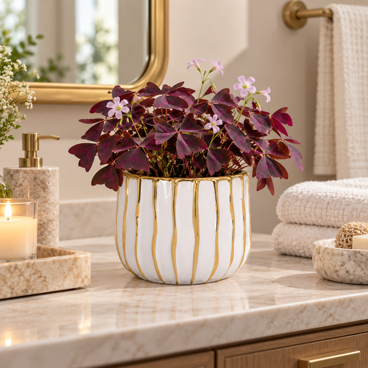 A potted ornamental plant with deep burgundy-purple leaves and small pale pink flowers sits on a marble bathroom counter in a real interior setting. The product appears to be the plant in a white ceramic pot with gold vertical stripes and gold drip detailing.