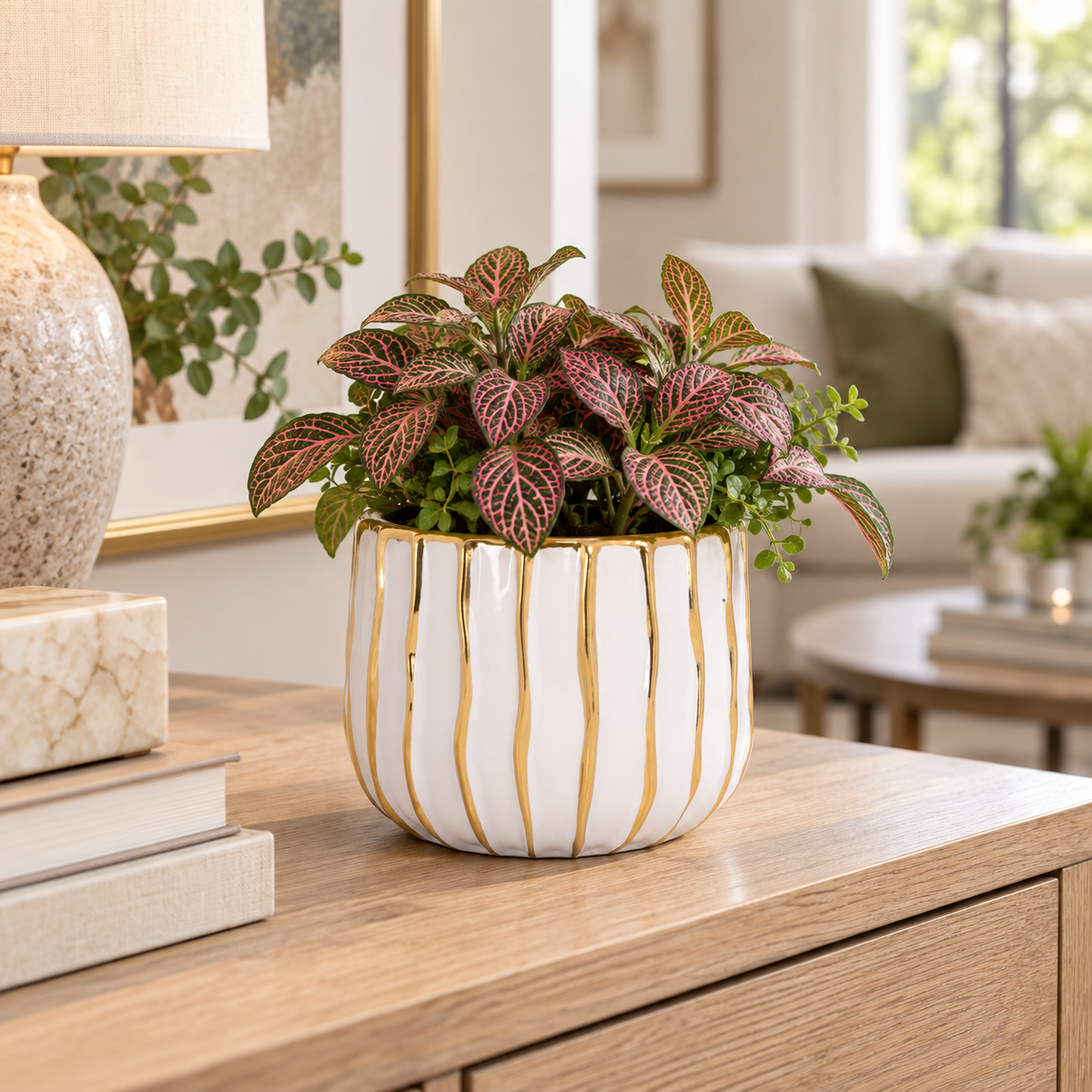 A potted houseplant with pink and green patterned leaves in a white ceramic pot with gold vertical stripes, displayed on a wooden console table in a bright, modern living room.