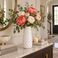 A white textured ceramic vase filled with a floral arrangement of coral peonies, blush roses, and green eucalyptus, styled on a marble console table in a bright, elegant hallway.