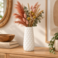 A white textured ceramic vase filled with dried decorative grasses, pink flower buds, and green eucalyptus leaves, displayed on a wooden console table in a styled interior setting.