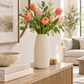 A tall matte off-white ceramic vase filled with pink tulips, peach tulips, and green eucalyptus stems, styled on a light wood console table in a bright, modern living room.