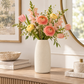 A tall matte off-white ceramic vase filled with a pastel floral arrangement featuring pink ranunculus, peach tulips, yellow flowers, and green foliage, styled on a wooden console table in a bright interior setting.