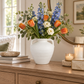 A white ceramic vase filled with a spring flower arrangement of orange tulips, blue delphiniums, and white roses, styled on a wooden console table in a warm, softly lit room.