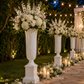 Outdoor wedding garden setup with tall white urn-style flower vases filled with white and blush floral arrangements, a stone aisle lined with candles and small flowers, warm string lights, greenery, and a draped white pavilion in the background.