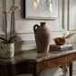 Two rustic terracotta clay jugs with handles displayed on a dark marble console table in a styled interior setting with a potted orchid, framed wall art, and books.
