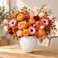 A vibrant autumnal flower bouquet in a textured light gray ceramic vase, set on a wooden table in a softly lit room.