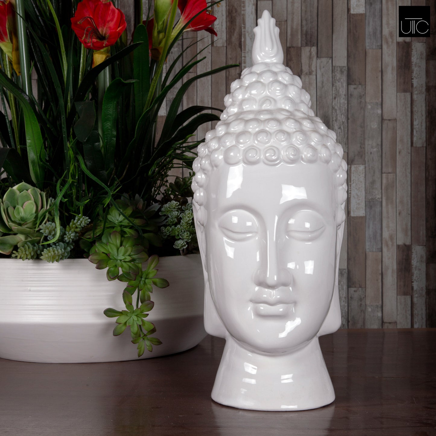 A white ceramic figurine of a Buddha head with a glossy finish, displayed in front of a wooden backdrop and accompanied by plants.