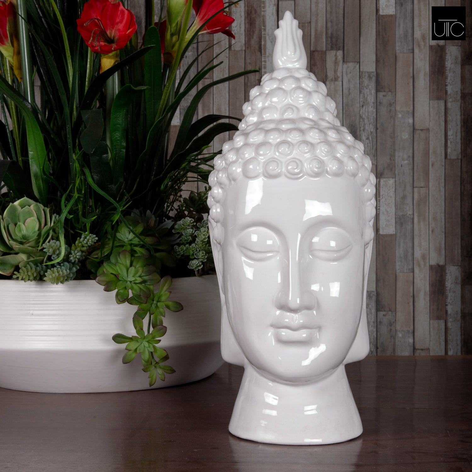 A white ceramic figurine of a Buddha head with a glossy finish, displayed in front of a wooden backdrop and accompanied by plants.