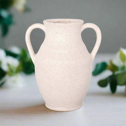 White ceramic vase with handles on a light surface with blurred greenery in the background