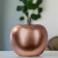 Bronze-colored apple-shaped object on a white surface with a blurred green plant in the background