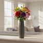 A tall ribbed gray vase holding a colorful bouquet of artificial flowers, placed on a light wood surface in a softly blurred living room setting.