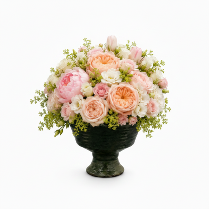 A floral arrangement of pink and peach roses, peonies, white blossoms, and green foliage in a dark green pedestal urn, photographed on a clean white background.