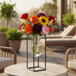 A vibrant artificial flower arrangement featuring red roses, orange and yellow sunflowers, purple peonies, pink carnations, and green foliage, displayed in a clear glass vase suspended within a black metal frame stand against a white background.