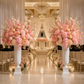 A grand wedding chapel aisle with two massive white pedestals holding towering floral arrangements in blush pink, peach, and cream tones. Rows of chairs line both sides under draped white curtains and hanging crystal chandeliers, with small candles placed along the floor.