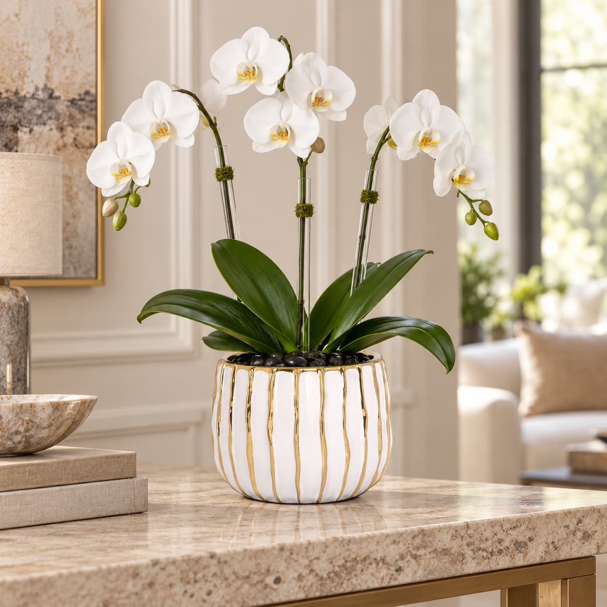 A potted white Phalaenopsis orchid with five blooms, displayed in a white ceramic pot with gold vertical stripes on a marble console table in a bright, elegant living room.
