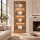 A tall wooden display cabinet with glass doors and warm interior lighting, styled with four white ceramic vases in a modern living room next to a sofa and fireplace.