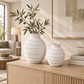 Two textured off-white ceramic vases with ribbed patterns, one large and one small, displayed on a light wood console table in a warm, modern living room setting.